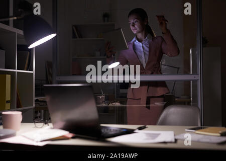 Young businesswoman standing looking at digital tablet in her hand and making notes on glass wall in dark office Stock Photo
