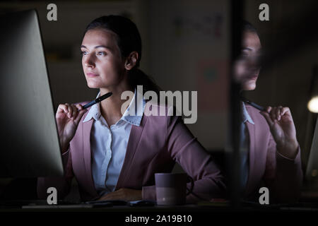 Concentrated young manager looking at computer monitor while working at her workplace till night at office Stock Photo