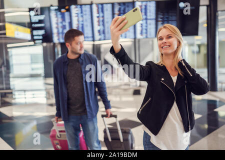 Image of man carrying luggage and woman taking selfie walking in waiting room at airport, blurred background. Stock Photo