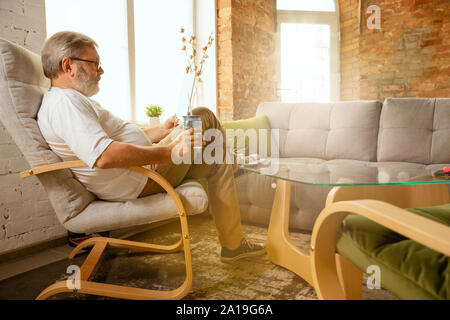 Senior man working with tablet at home - concept of home studying. Caucasian male model sitting on sofa and drinking water while serfing in internet, watching cinema or webinar, online lessons. Stock Photo