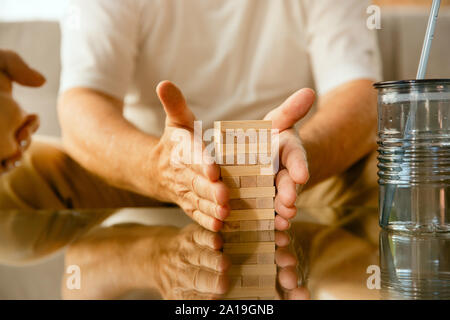 Close up of senior man's hands doing his wooden constructor at home - concept of home studying. Caucasian male model sitting on sofa and working with bricks for training of fine motor skills. Stock Photo
