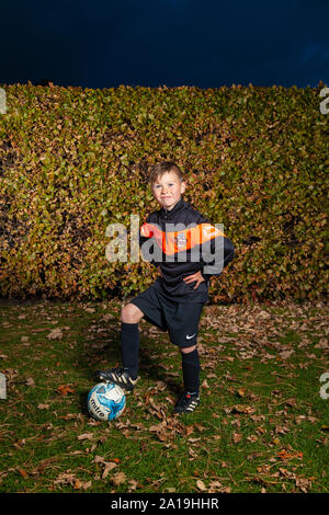 An eight year old boy wearing a football strip with arms on hips and his foot on a football Stock Photo