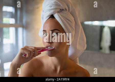 Young brunette woman brushing her teeth in a bathroom with her hair wrapped in a towel Stock Photo