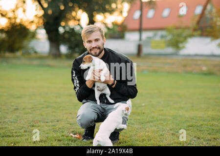 Young man sits at lawn and plays with Jack Russell puppies. Stock Photo