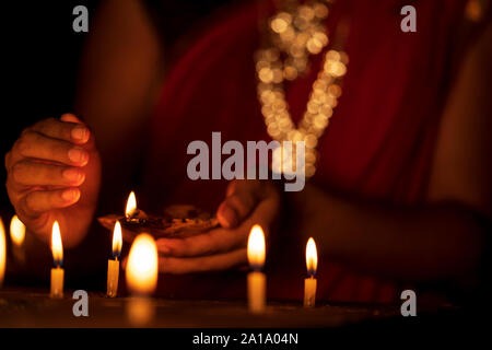 Indian housewife or newly bride woman wearing traditional red saree & gold jewelry lighting candles with a clay diya or oil lamp in one hand at a temp Stock Photo