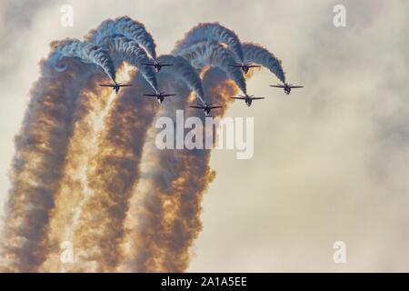 The British RAF red arrows display team in a seven ship close formation rolling inverted trailing plumes of smoke against a sunlit sky Stock Photo