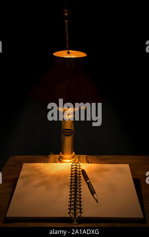 Dark, moody image of an open journal with a pencil on a wooden table, lit by an old weathered table lamp; focus on pencil Stock Photo