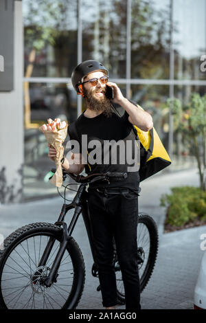 Young and cheerful courier waiting with flowers for a client near the building outdoors, delivering fresh floristry on a bicycle using thermal bag Stock Photo
