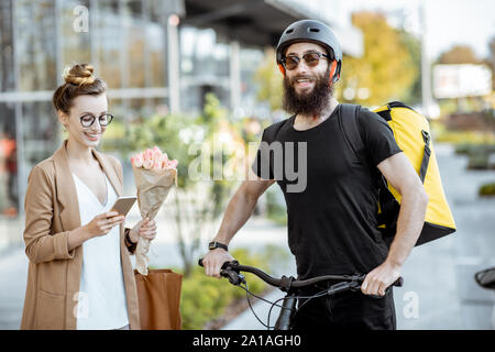 Courier delivering fresh flowers to a young business woman on a bicycle wearing delivery backpack. Fresh floral delivery concept Stock Photo