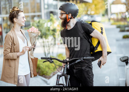 Courier delivering fresh flowers to a young business woman on a bicycle wearing delivery backpack. Fresh floral delivery concept Stock Photo