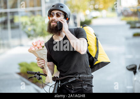 Young and cheerful courier waiting with flowers for a client near the building outdoors, delivering fresh floristry on a bicycle using thermal bag Stock Photo