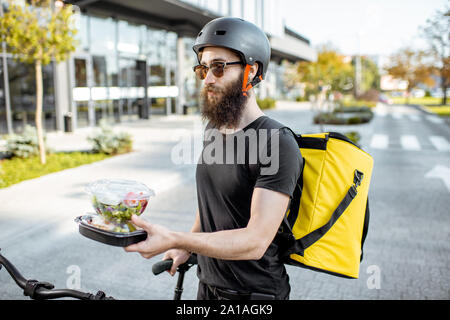 Young and cheerful courier waiting for a client with takeaway lunches near the building outdoors, delivering restaurant food on a bicycle using therma Stock Photo
