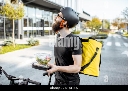 Young and cheerful courier waiting for a client with takeaway lunches near the building outdoors, delivering restaurant food on a bicycle using therma Stock Photo
