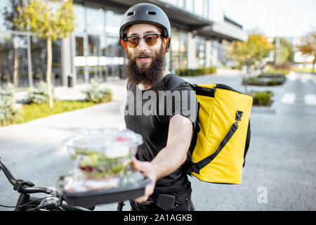 Portrait of a young and cheerful courier giving takeaway lunches, delivering restaurant food on a bicycle using thermal bag Stock Photo