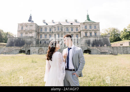 Fabulous Asian wedding couple posing in front of an old medieval castle, hugging and kissing on a sunny day. Stock Photo