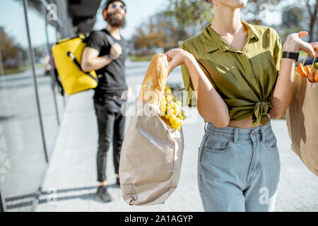 Woman carrying paper bags full of fresh food received from a courier. Close-up view focused on products with delivery man on the background Stock Photo