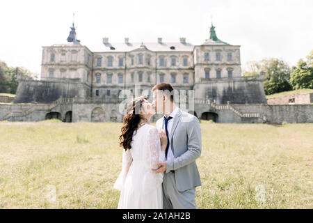 Fabulous Asian wedding couple posing in front of an old medieval castle, hugging and kissing on a sunny day. Stock Photo