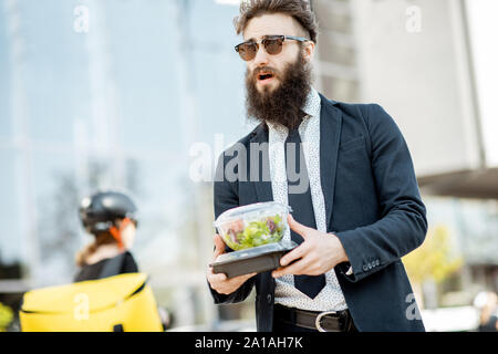 Elegant bearded office worker carrying takeaway lunch received from a courier near the office building. Female courier with delivery bag on the backgr Stock Photo