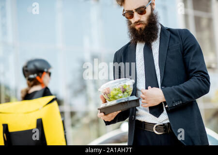 Elegant bearded office worker carrying takeaway lunch received from a courier near the office building. Female courier with delivery bag on the backgr Stock Photo