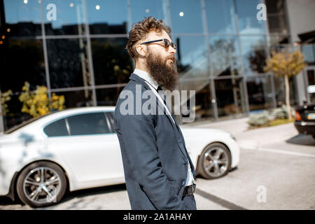 Portrait of an elegant and confident bearded businessman dressed in the suit walking near the car and office building outdoors Stock Photo
