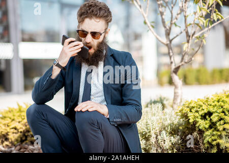 Elegant bearded businessman dressed in the suit having emotional talk using smartphone while sitting near the office outdoors Stock Photo