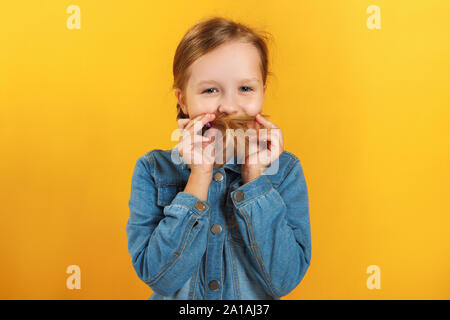 Cheerful happy little girl makes a mustache from her hair. A child in a denim shirt on a yellow background. Stock Photo