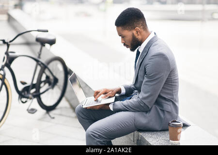 Serious businessman using laptop outdoors with bike nearby Stock Photo