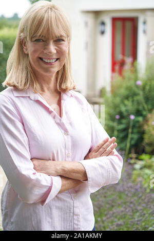 Portrait Of Mature Woman Standing In Garden In Front Of Dream Home In Countryside Stock Photo
