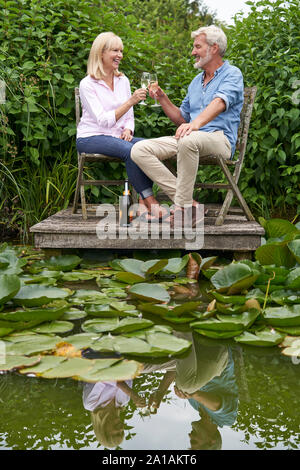 Mature Couple Celebrating With Champagne Sitting On Chairs On Wooden Jetty By Lake Stock Photo
