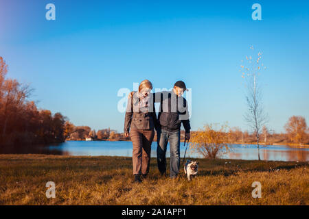 Senior couple walking pug dog in autumn park by river. Happy man and woman enjoying time with pet. Stock Photo