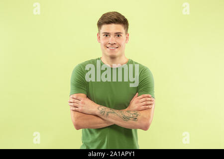 Caucasian young man's half-length portrait on green studio background. Beautiful male model in shirt. Concept of human emotions, facial expression, sales, ad. Standing with hands crossed, smiling. Stock Photo