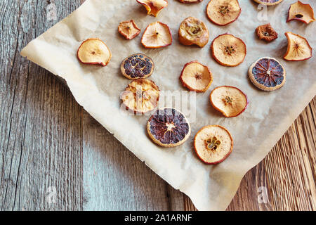 Homemade slices of dried oranges and apples on baking paper and black baking tray, diagonal arrangement from top right corner, wooden background Stock Photo