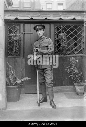 A vintage black and white photograph showing a young man, wearing spectacles dressed in a First World War British Army soldier, uniform, standing in the back yard of a house, leaning on his rifle.