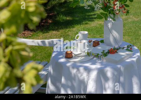 Enjoying coffee time on a sunny spring day. Served table with coffee, canele and flowers in the garden. There is a white wooden bench next to it Stock Photo