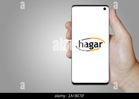 A man holds a smartphone displaying the logo of company Hagar listed on Nasdaq Iceland. Grey gradient in the background. Credit: PIXDUCE Stock Photo