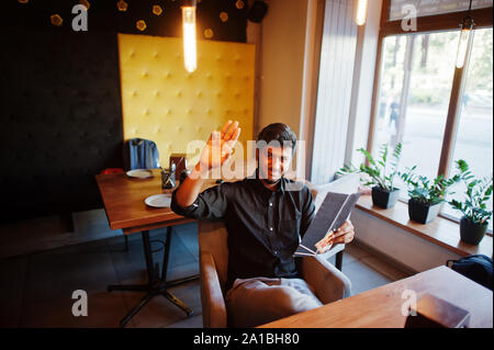 Confident young indian man in black shirt sitting at cafe, read menu and calling waiter. Stock Photo