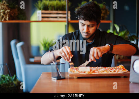 Confident young indian man in black shirt sitting at pizzeria with pizza.