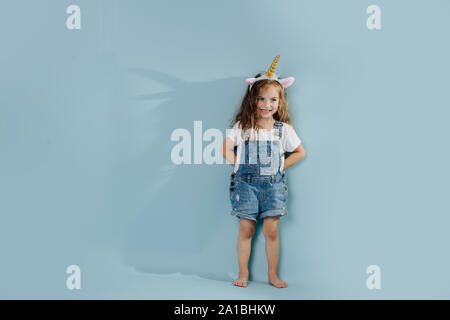 Little girl, wearing unicorn headband, leaning against blue wall. Stock Photo