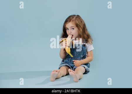 Little girl is eating banana, while sitting on the floor over blue background Stock Photo