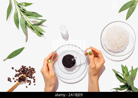 Woman's hands adding oil using pipette into ground coffee leftovers to make homemade natural body scrub on white table, flat lay composition. Stock Photo