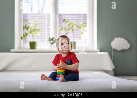 Happy kid with coloured pyramid toy on white bed in his room