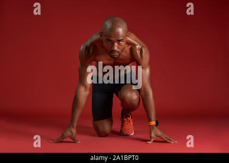 Portrait of young african man in sportswear going to run while sitting isolated over red background. Fitness and work out concept. Horizontal shot Stock Photo