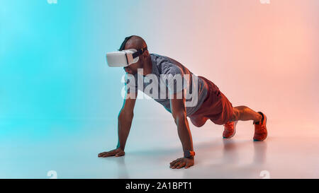 VR technology. Full length of young african man in sports clothing doing plank while wearing virtual reality glasses. Studio shot. Future technology Stock Photo