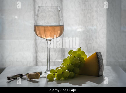 Glass of rose wine on the table, next to green grapes, hard cheese, corkscrew with cork. Close-up, backlight Stock Photo