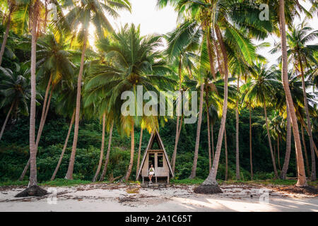 Yung woman sits in doorway of hut and holds coconut in her hands. Jungle wildlife in Port Barton, Philippines Stock Photo