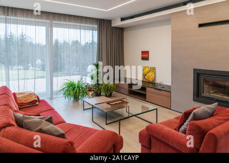 Living room with sofa and table. To the right is the fireplace. There are flowers in front and a window overlooking the terrace and garden Stock Photo