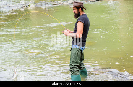 Fun of fishing is catching. Fishing masculine hobby. Brutal man wear rubber  boots stand in river water. Fisher weekend activity. Fisher with fishing  equipment. Fish on hook. Leisure in wild nature Stock
