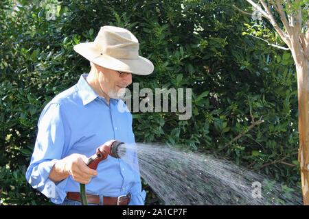 Watering the plants, senior man wearing a hat in his garden in the early morning light Stock Photo