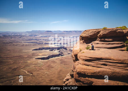 Mid aged caucasian man is sitting at the edge of the cliff and looking away at the valley among red rocks of the canyons Stock Photo