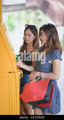 Two young asian female withdrawing the money from a bank card using ATM machine at the mall Stock Photo